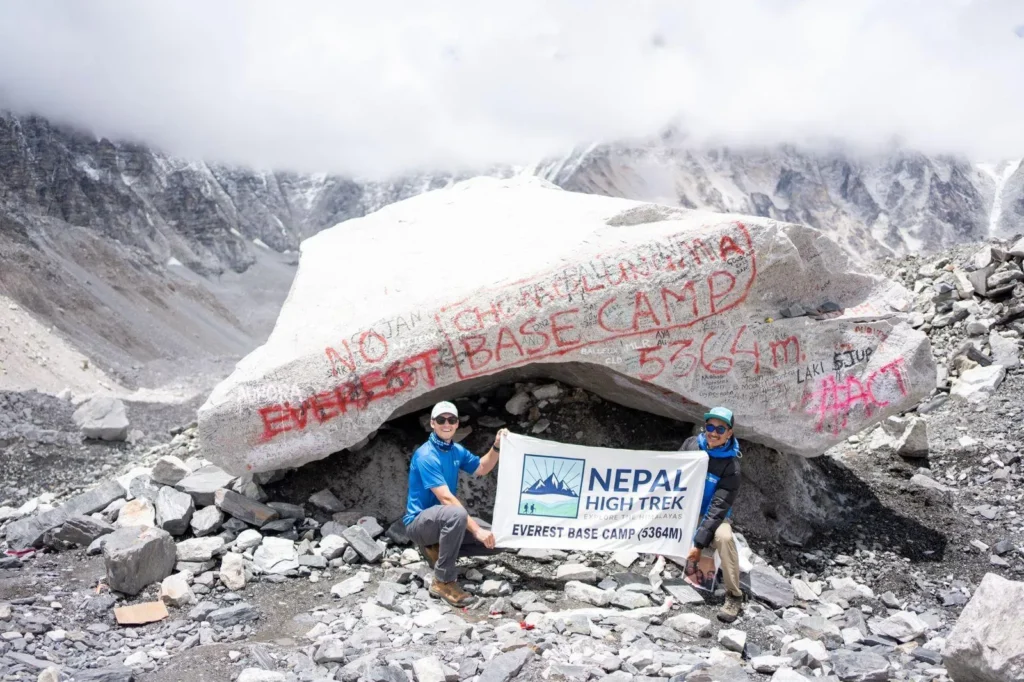 Two smiling trekkers holding a 'Nepal High Trek' banner at the base of a large, graffiti-covered rock marking Everest Base Camp (5364m), with rocky mountains in the background.