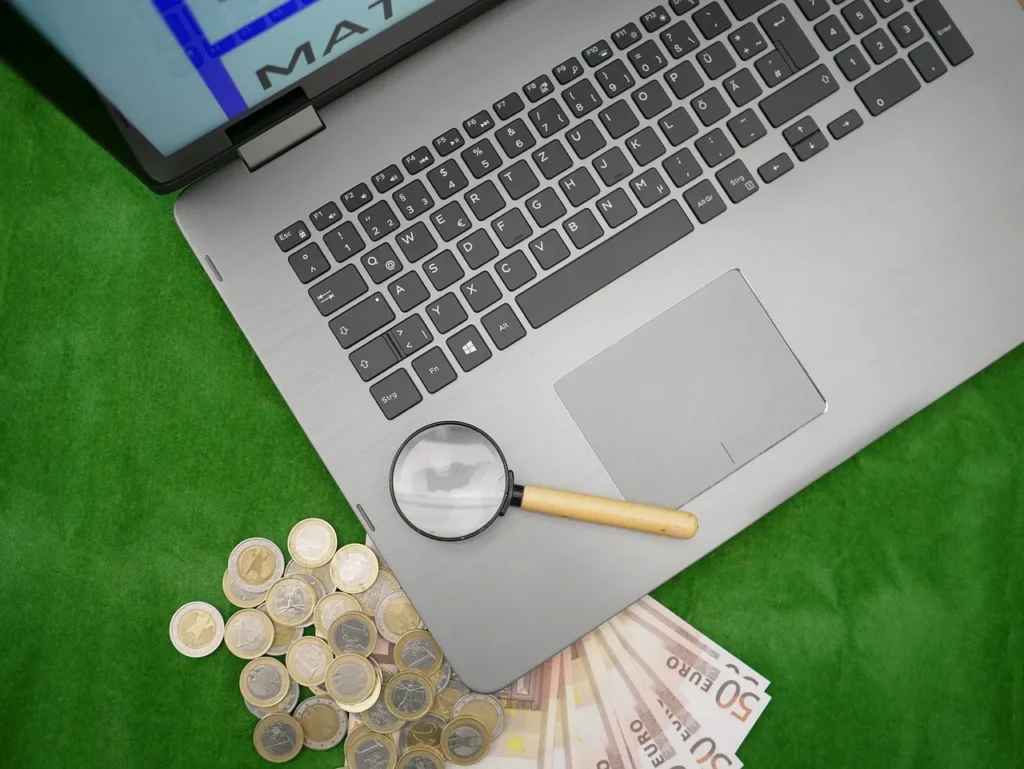 A magnifying glass lies on a laptop keyboard, next to scattered Euro coins and banknotes, all on a green felt surface, symbolizing the scrutiny of online betting finances.