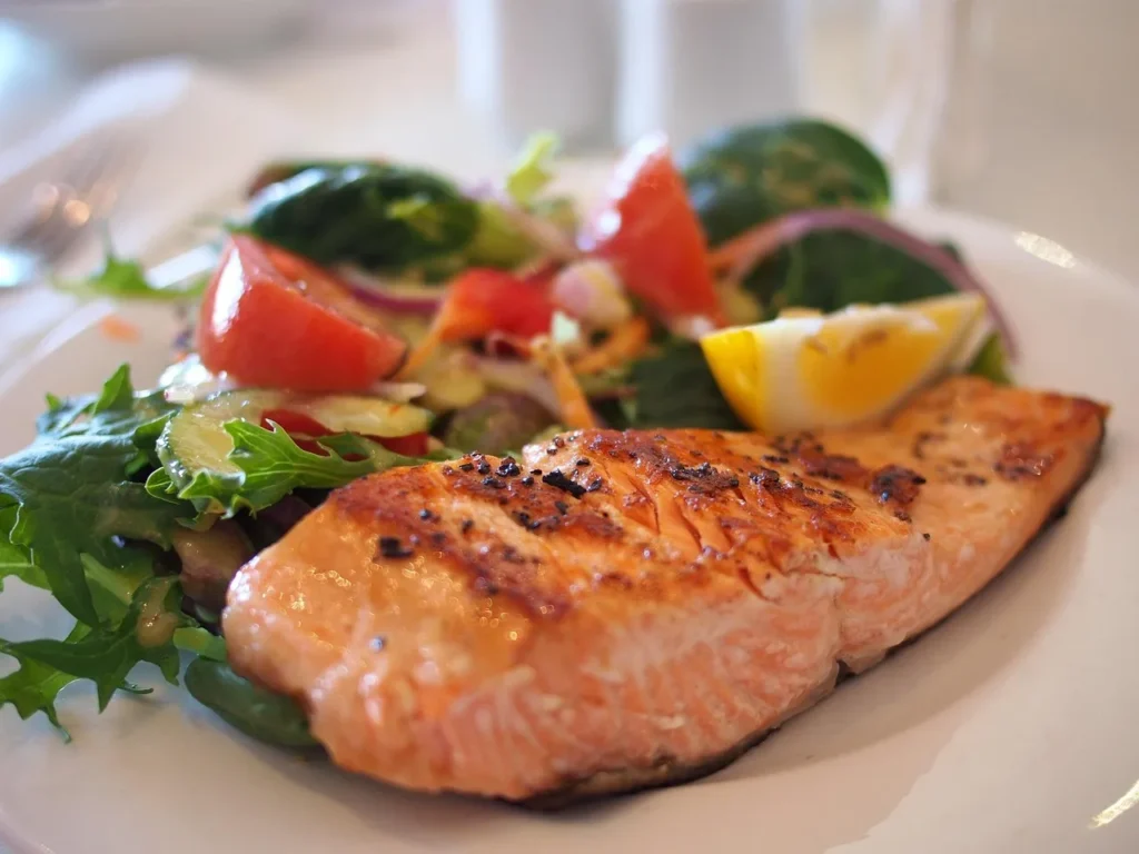A grilled salmon fillet seasoned with pepper, served alongside a fresh green salad with tomatoes, red onion, and a lemon wedge, exemplifying a healthy Mediterranean-style meal.