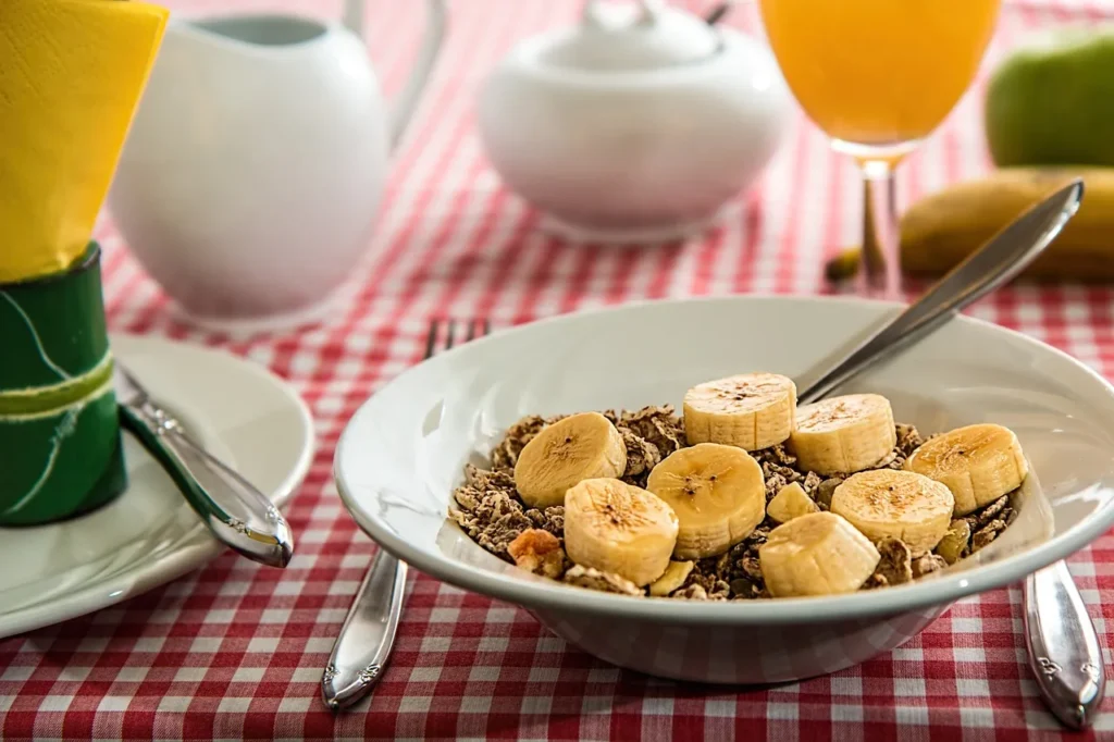 A bowl of healthy breakfast cereal topped with sliced bananas, sitting on a red and white checkered tablecloth, with a glass of orange juice and other breakfast items blurred in the background.