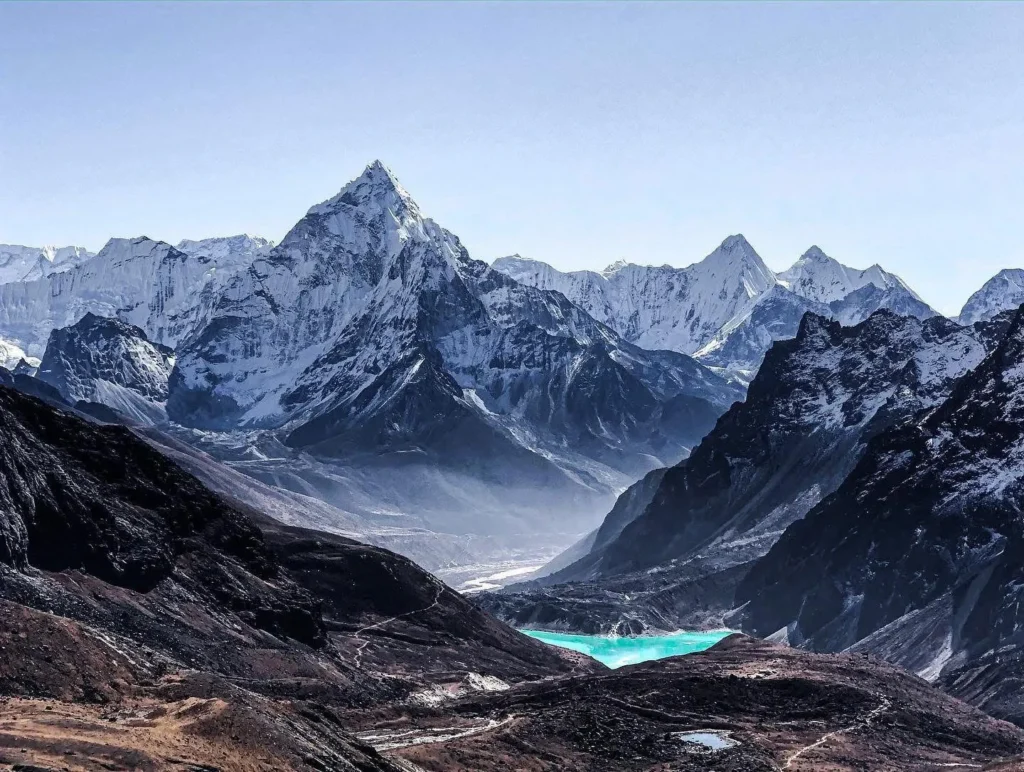A stunning panoramic view of the Himalayas featuring snow-capped peaks, including the prominent Ama Dablam, with a vibrant turquoise glacial lake nestled in the foreground valley.