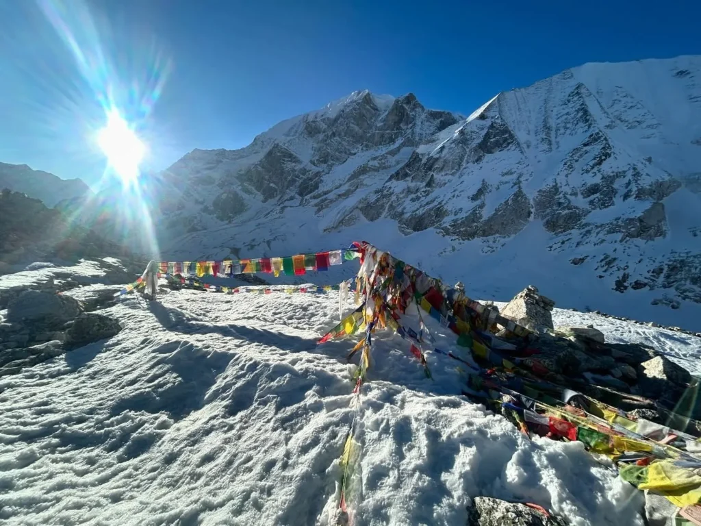 Colorful prayer flags strung across a snow-covered mountain landscape, with towering, white peaks under a bright sunburst in the background.