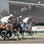 Three jockeys on horseback race neck-and-neck on a dirt track, with a large screen in the background showing another horse race in progress.
