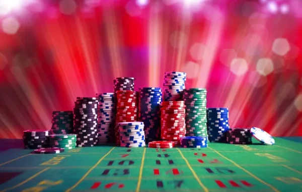A vibrant image of numerous stacks of colorful casino chips (red, blue, green, black, white) on a green felt roulette table, with a blurred background of red and purple light streaks, suggesting an exciting casino atmosphere.