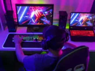 A person from behind, wearing headphones and a baseball cap, intensely plays video games on a brightly lit desktop computer setup with a second laptop visible to the right. The room is bathed in vibrant neon pink and blue lighting.