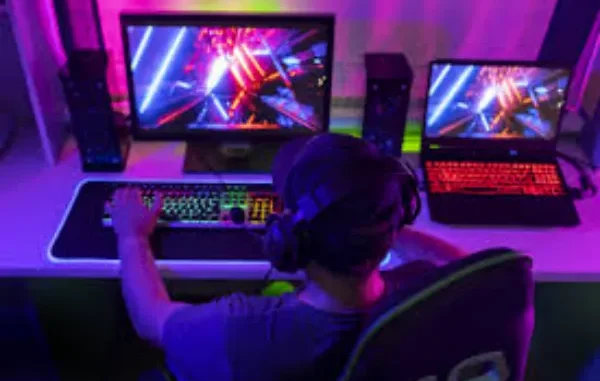 The Future of Immersive Gaming: A Glimpse into High-Tech Setups A person from behind, wearing headphones and a baseball cap, intensely plays video games on a brightly lit desktop computer setup with a second laptop visible to the right. The room is bathed in vibrant neon pink and blue lighting.