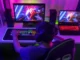 A person from behind, wearing headphones and a baseball cap, intensely plays video games on a brightly lit desktop computer setup with a second laptop visible to the right. The room is bathed in vibrant neon pink and blue lighting.