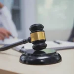 A wooden gavel rests on a desk in the foreground, with blurred figures of people in professional attire discussing documents in the background, symbolizing legal proceedings or a court of law.