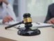 A wooden gavel rests on a desk in the foreground, with blurred figures of people in professional attire discussing documents in the background, symbolizing legal proceedings or a court of law.