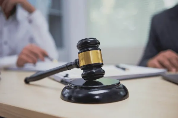 A close-up of a wooden gavel on a light-colored desk, with two blurred individuals in the background reviewing documents, suggesting a legal consultation or discussion related to personal injury law.