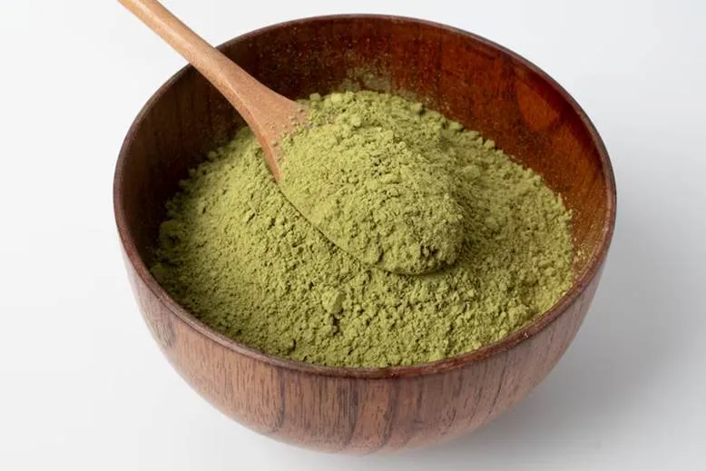 A wooden bowl filled with vibrant green kratom powder, with a light wooden spoon resting in it, on a white background.