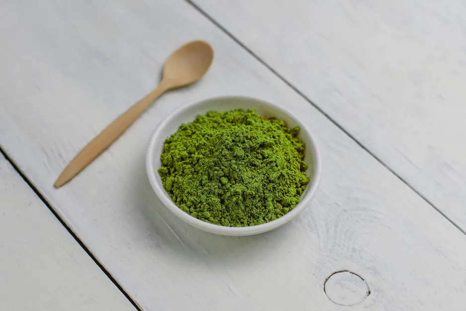 A white bowl filled with vibrant green kratom powder, next to a light wooden spoon on a white wooden surface.