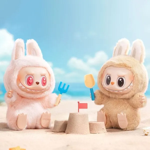 wo plush Labubu figures, one pink and one brown, sitting on a sandy beach with a small sandcastle, holding miniature beach toys.