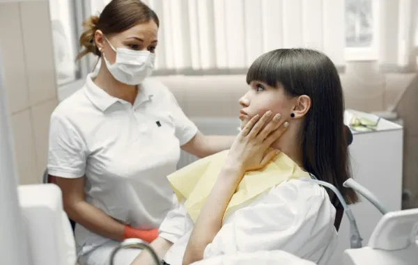 A young woman with dark hair, wearing a white shirt and a yellow dental bib, sits in a dentist's chair, holding her jaw with a pained expression. A female dental professional, wearing a white polo shirt, face mask, and orange gloves, stands beside her, looking at the patient with concern.