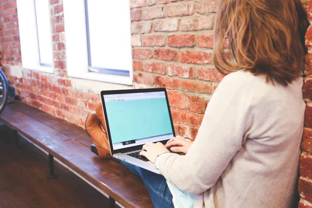 A person, seen from behind, sits on a wooden bench with their laptop open, typing on the keyboard. They are casually dressed and are positioned against a rustic brick wall, suggesting a humble beginning or a focus on work regardless of environment. The laptop screen is a light teal, and a window is visible to the left.