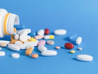 A white prescription bottle on its side with a variety of colorful pills and capsules spilled out onto a vibrant blue surface.