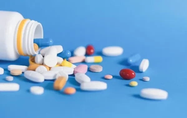 A white prescription bottle on its side with a variety of colorful pills and capsules spilled out onto a vibrant blue surface.