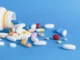 A white prescription bottle on its side with a variety of colorful pills and capsules spilled out onto a vibrant blue surface.