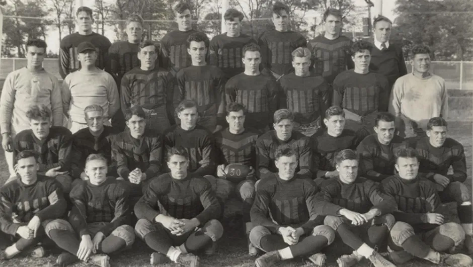 Black and white group portrait of the 1930s NC State (then State College) football team, consisting of a large number of players in dark, padded uniforms, posing in rows on a grass field.