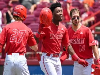 Three NC State Wolfpack baseball players in red jerseys celebrating on the field, with the central player holding his helmet aloft, conveying excitement and team spirit after a victory.