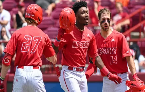 NC State Baseball: A Legacy of Victory and Passion Three NC State Wolfpack baseball players in red jerseys celebrating on the field, with the central player holding his helmet aloft, conveying excitement and team spirit after a victory.