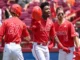 Three NC State Wolfpack baseball players in red jerseys celebrating on the field, with the central player holding his helmet aloft, conveying excitement and team spirit after a victory.