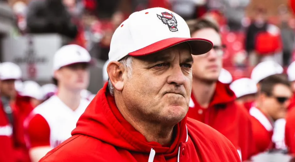 Close-up of NC State Baseball Head Coach Elliott Avent wearing a red hoodie and a white and red NC State Wolfpack baseball cap, with a focused expression, likely on the field during a game.