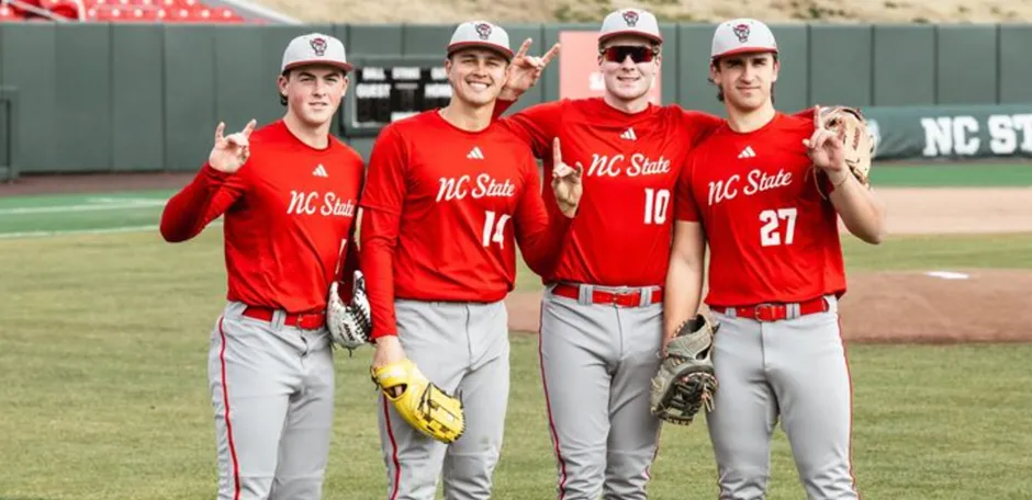 Four NC State Wolfpack baseball players in red jerseys, grey pants, and caps posing together on the field, with some making hand gestures like the "Wolfpack" sign, showcasing team camaraderie.