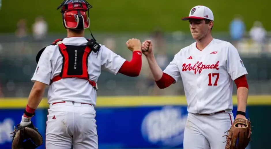 An NC State Wolfpack baseball pitcher (number 17, wearing a white uniform with "Wolfpack") and catcher (wearing white with red gear) exchange a fist bump on the field, showing team synergy and focus during a game.