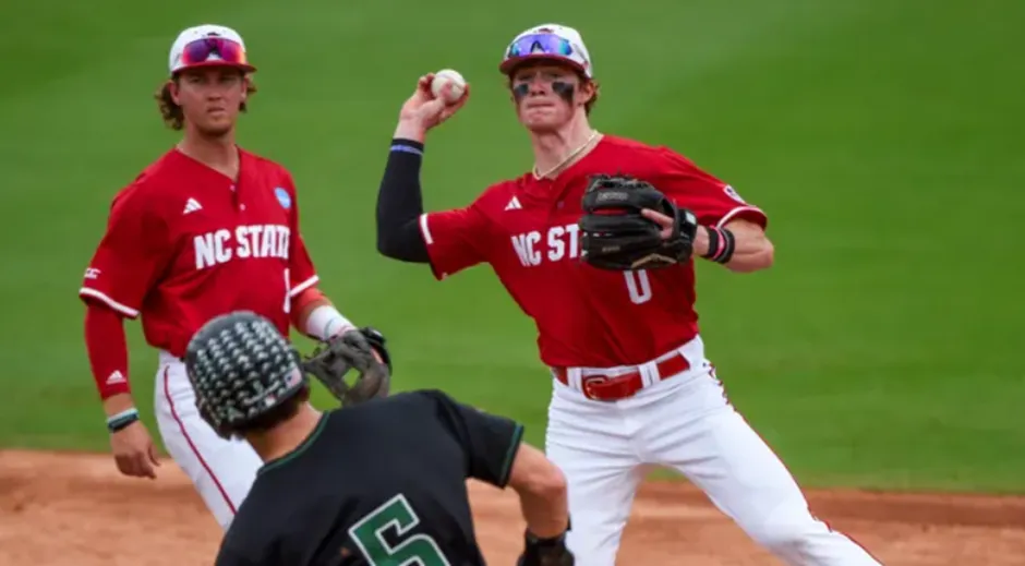An NC State Wolfpack baseball player in a red jersey and white pants throws a baseball during a game, with another Wolfpack player observing and an opposing player's back visible in the foreground.