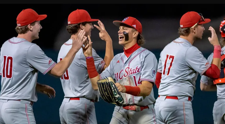 Four NC State Wolfpack baseball players in gray and red uniforms celebrating enthusiastically on the field, with one player showing a wide smile, eye black, and gloved hand, receiving high-fives from teammates.