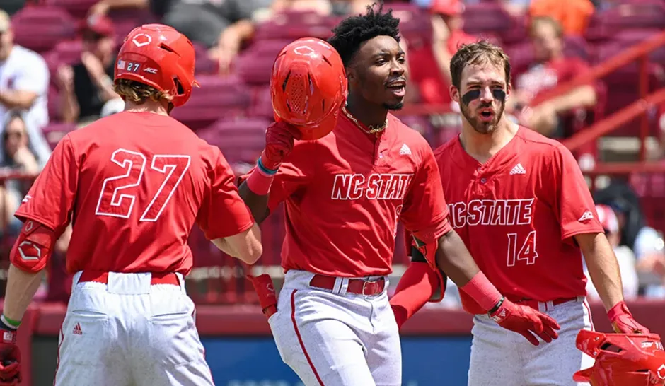 Three NC State Wolfpack baseball players in red uniforms celebrating on the field, with one player holding his red helmet up, depicting a moment of shared victory and excitement.