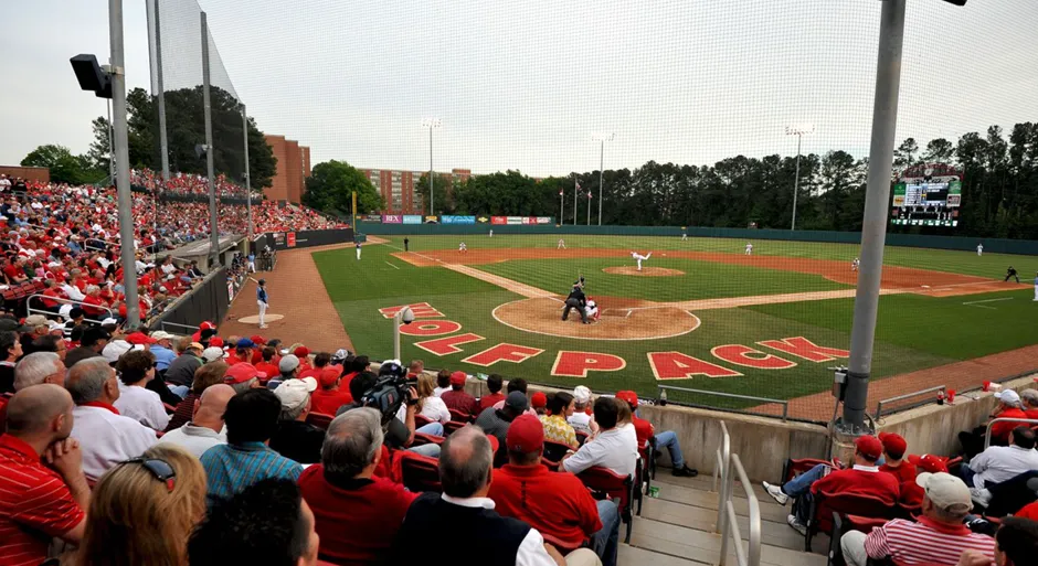 Panoramic view of a baseball game in progress at Doak Field, NC State's baseball stadium, showing players on the field, a batter at home plate, and fans in the stands, under a clear sky.
