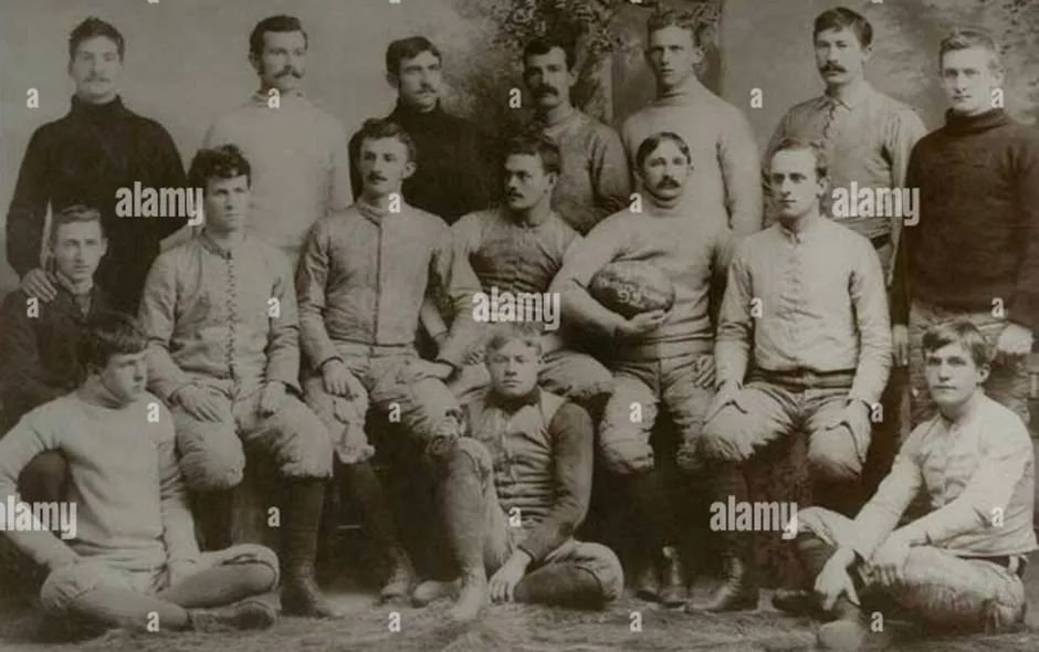 Sepia-toned photograph of the very first North Carolina A&M (later NC State) football team, a group of 17 young men in early football uniforms posing formally, with some seated and others standing, one holding a vintage football.