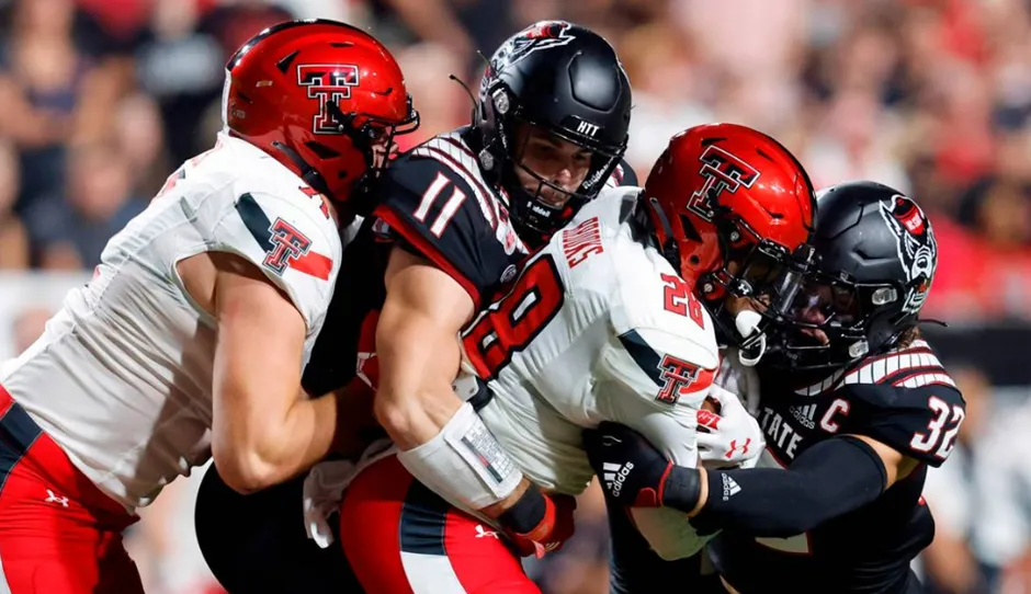Intense college football action shot featuring NC State Wolfpack players in black uniforms with red accents tackling a Texas Tech Red Raiders player in a white and red uniform, surrounded by other players.