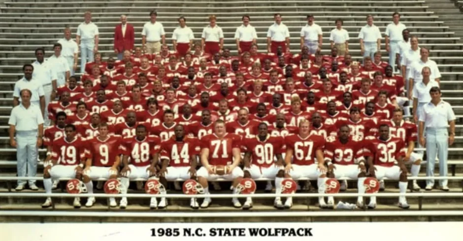 Large group photograph of the 1985 NC State Wolfpack football team, featuring numerous players in red jerseys with white numbers, coaches, and staff members, all posing on stadium bleachers with helmets lined up in front of the players.