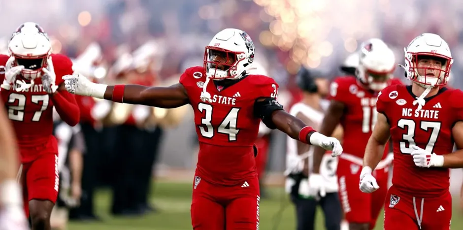 An NC State Wolfpack football player in a red uniform with the number 34 celebrates with arms outstretched on the field, surrounded by teammates and a blurred background suggesting a stadium atmosphere with pyrotechnics or smoke.