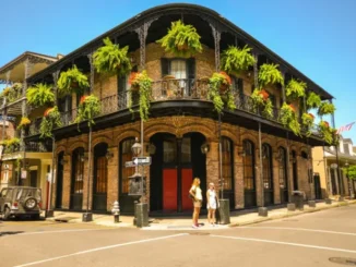 A historic brick building with wrought iron balconies adorned with hanging ferns and flowers in New Orleans, with two people standing on the sidewalk.