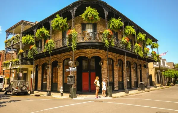 A historic brick building with wrought iron balconies adorned with hanging ferns and flowers in New Orleans, with two people standing on the sidewalk.