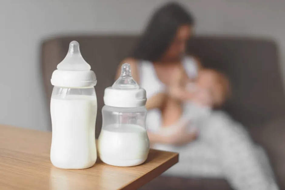 Two baby bottles filled with milk sit on a wooden table in the foreground, while a blurred image of a mother breastfeeds her baby on a couch in the background.