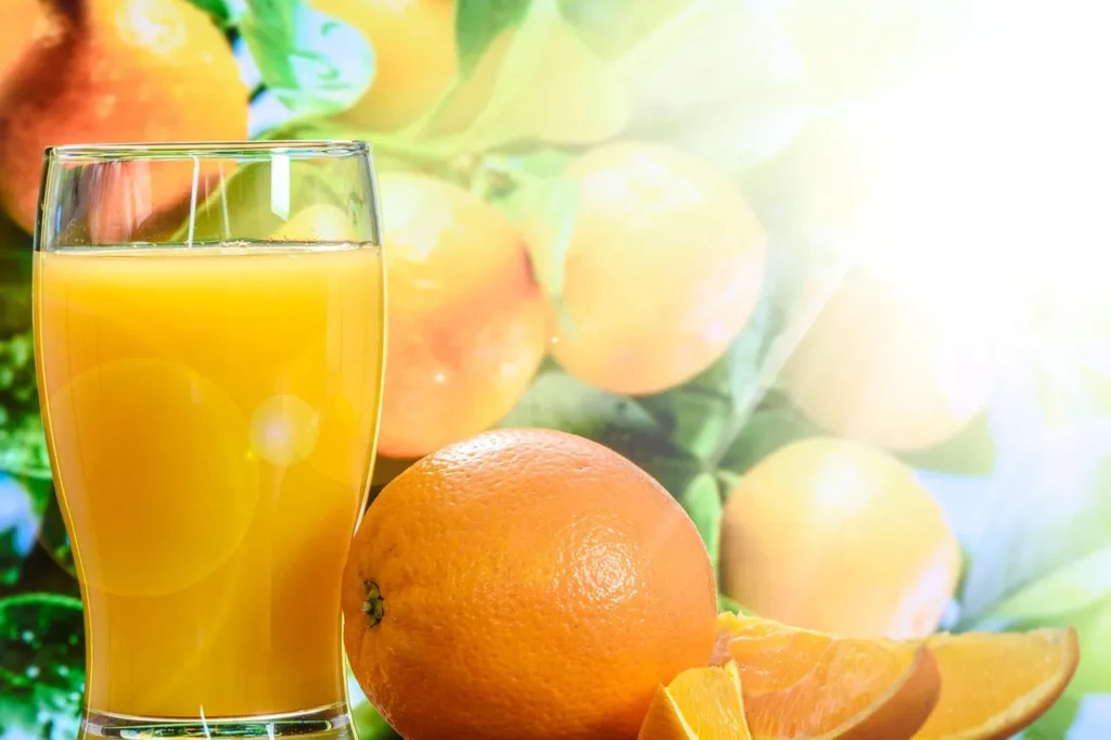 A glass of fresh orange juice next to a whole orange and orange slices, with blurred oranges on a tree in the background, highlighting the importance of vitamin C and hydration.