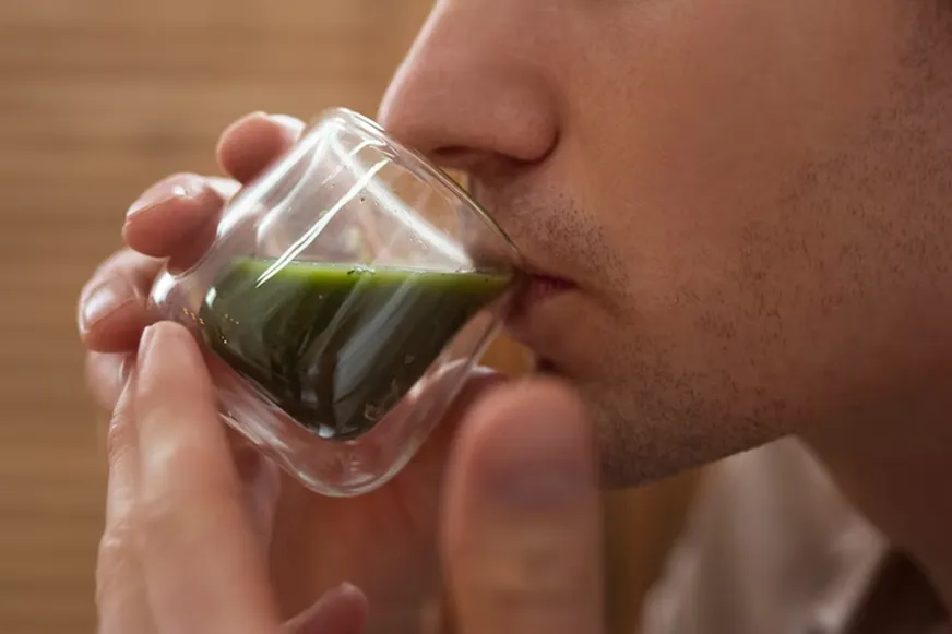 A close-up of a person drinking a green liquid from a small, clear glass, suggesting the consumption of a natural supplement like kratom.