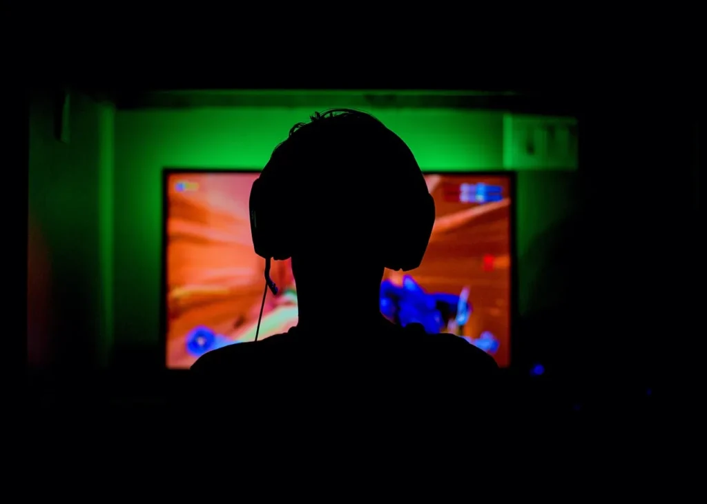 Silhouette of a person wearing gaming headphones, focused on a brightly lit screen displaying a video game, symbolizing intense gaming and concentration on competitive settings.