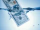 A hundred-dollar bill partially submerged in clear blue water, with ripples and small splashes.