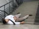 A man in a white shirt and khaki pants lying injured on the steps of a concrete staircase after a fall.