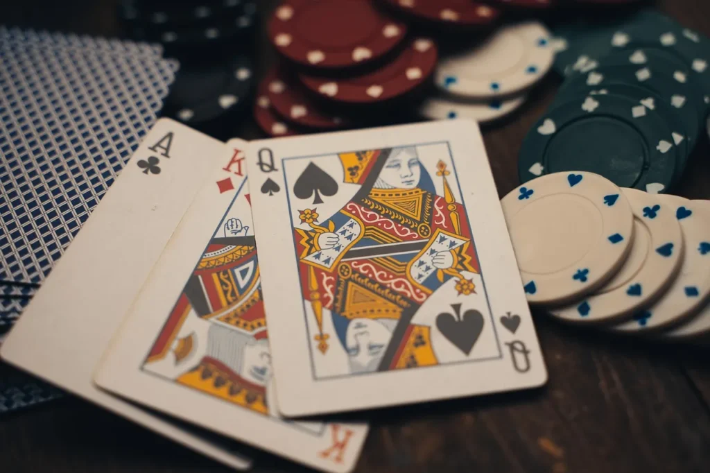 A pair of playing cards, an Ace of Spades and a Queen of Spades, laid out on a wooden surface next to stacks of poker chips in various colors, with a fanned deck visible in the background.