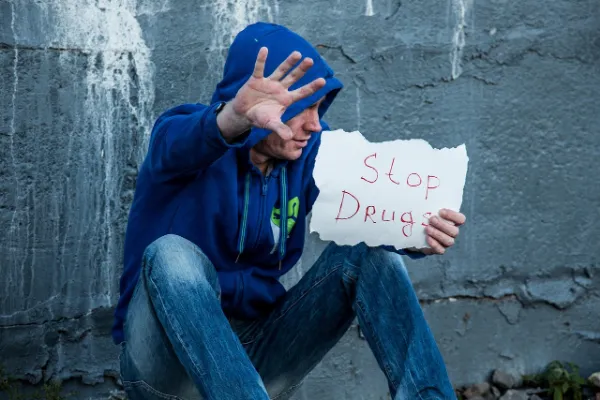 A person in a blue hoodie and jeans sitting against a distressed concrete wall, holding a crumpled piece of paper with "Stop Drugs" written in red, with one hand raised as if to push something away.