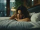 A young woman with wavy brown hair lies awake on a bed, propped on her forearms, with a thoughtful or concerned expression, illuminated by soft, dim light.