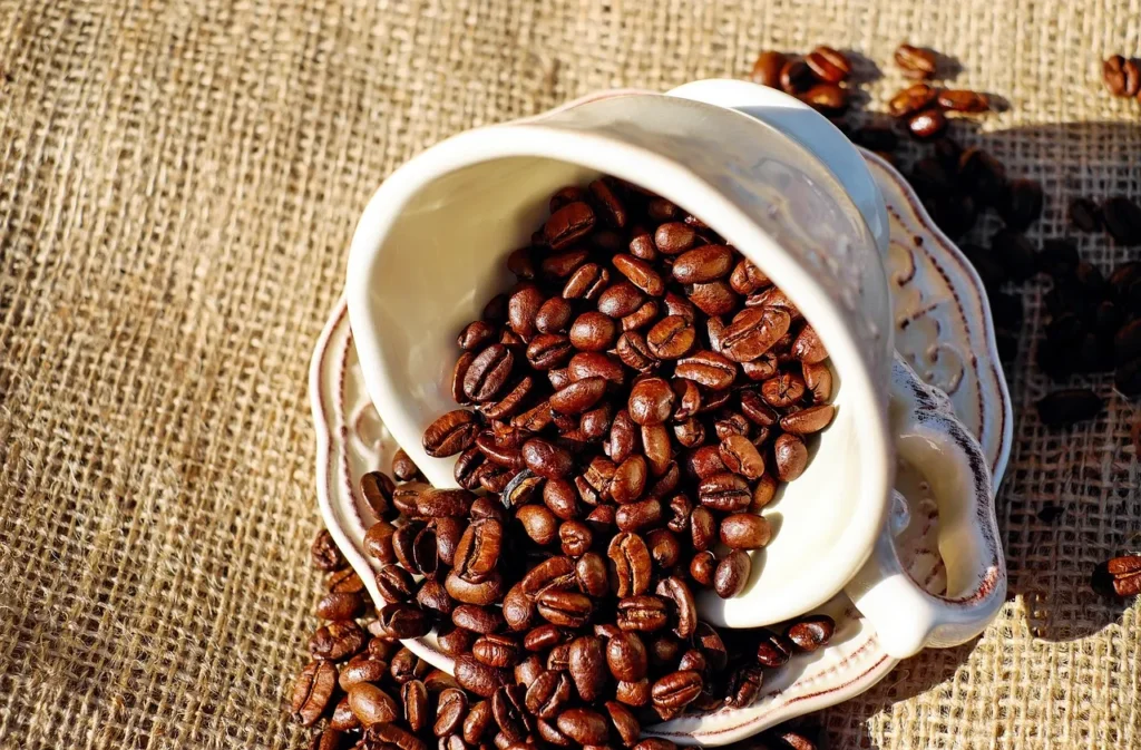 Dark roasted coffee beans spilling out of a decorative white ceramic cup and saucer, resting on a rustic burlap surface in sunlight in Dubai.