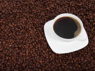 A white coffee cup filled with black coffee, set on a white saucer, surrounded by a large quantity of dark roasted coffee beans.